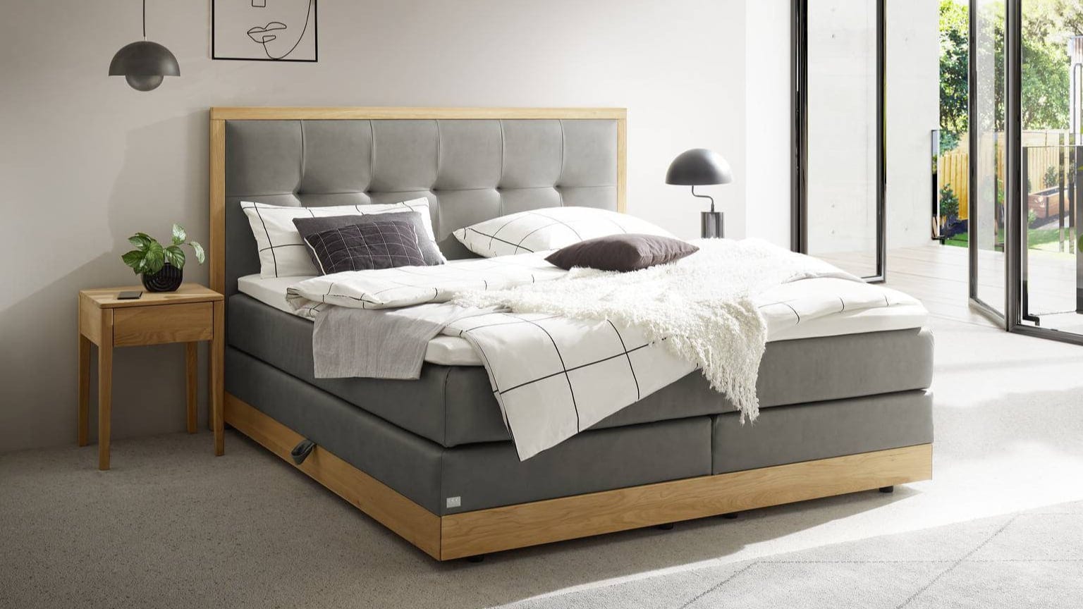 Modernes Schlafzimmer mit einem Boxspringbett von Hasena mit grauem Polsterkopfteil, weißer und grauer Bettwäsche, einem Holzrahmen, einem Nachttisch mit einer Pflanze und großen Fenstern, die natürliches Licht hereinlassen.
